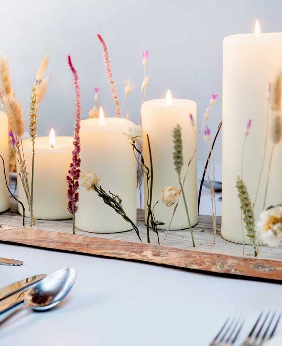 Elfenbeinfarbene Stumpenkerzen in verschiedenen Größen, arrangiert auf einem groben Holzbrett, das mit bunten, getrockneten Blüten besetzt wurde. Das Brett steht auf einem weißen, gedeckten Tisch zwischen dem Geschirr.
