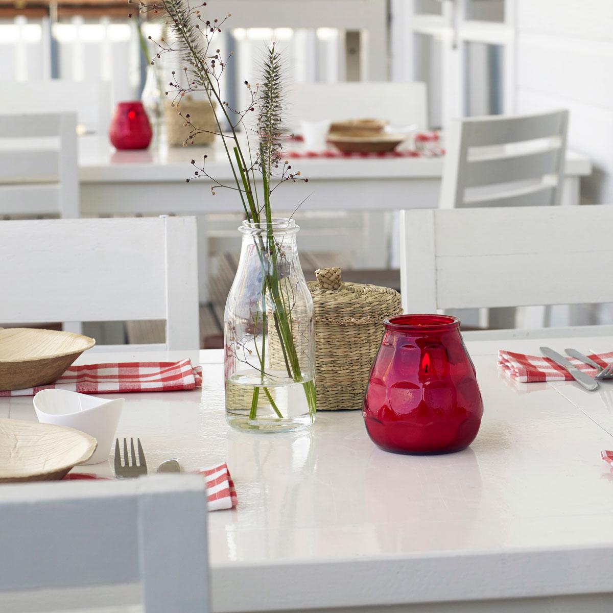 Tischdeko für die Gastronomie im Außenbereich: Weiße Holzmöbel mit sommerlich dekorierten Tischen. Rot-weiß karierte Servietten als Unterlage für kleine Holzschüsseln, geflochtene Körbchen aus Seegras und Glasvasen mit sommerlichen Zweigen. In der Mitte jedes Tisches steht ein rotes, brennendes Glaswindlicht.