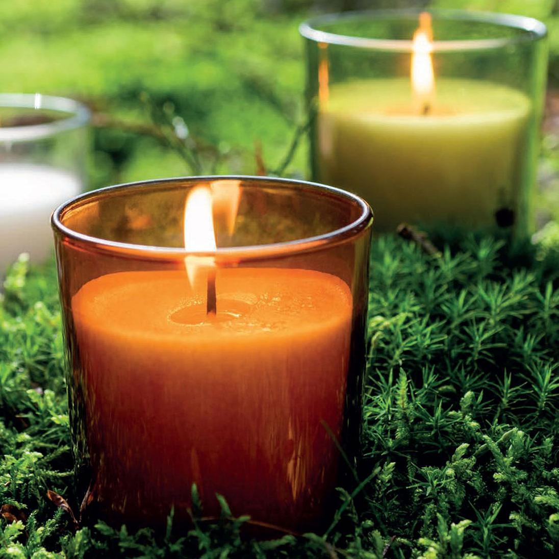 Nahaufnahme einer brennenden Duftkerze in einem orangefarbenen Glas. Im Hintergrund erkennt man noch zwei weitere Kerzen, eine mit grünem Glas und eine mit klarem. Die Gläser stehen auf dichtem, grünem Moos.