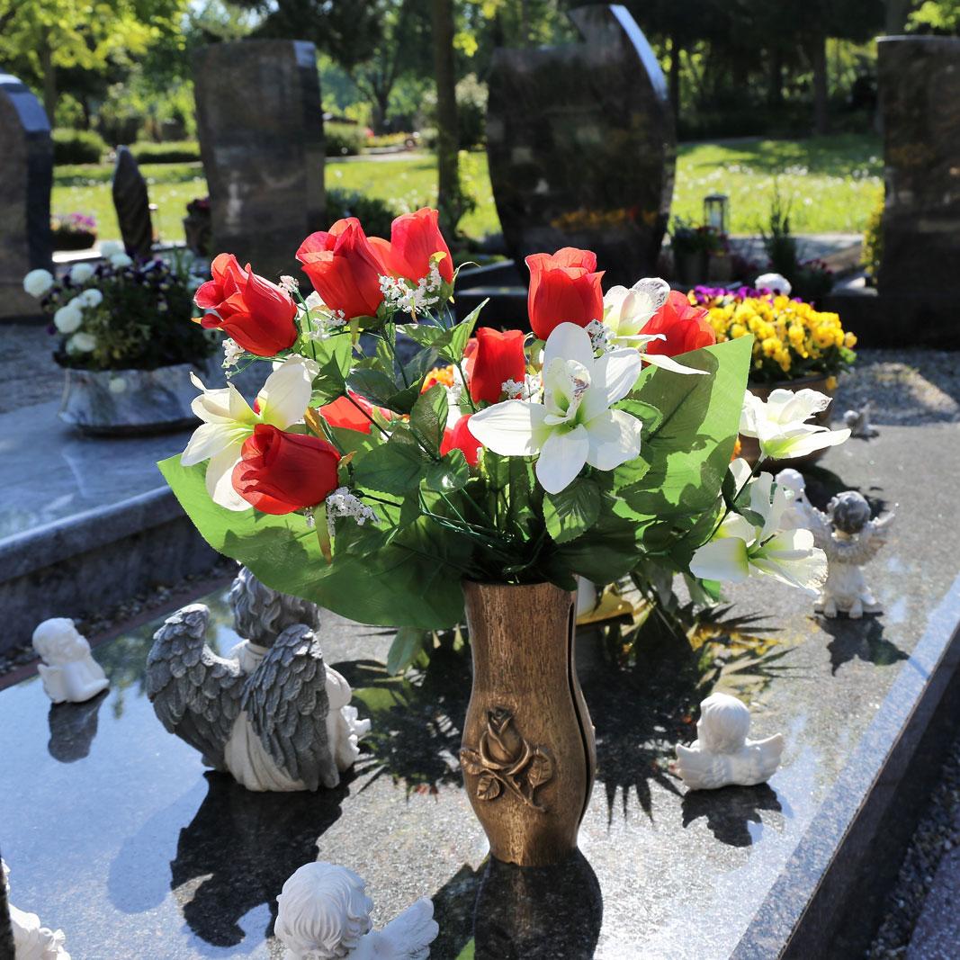 Grabstätte mit einer schwarzen Marmorplatte, auf der verschiedene Arten von Grabschmuck stehen. In einer bronzenen Grabvase steckt ein Blumenstrauß aus roten Rosen und weißen Lilien. Über das restliche Grab wurden kleine Grabengel in verschiedenen Größen und Positionen verteilt.