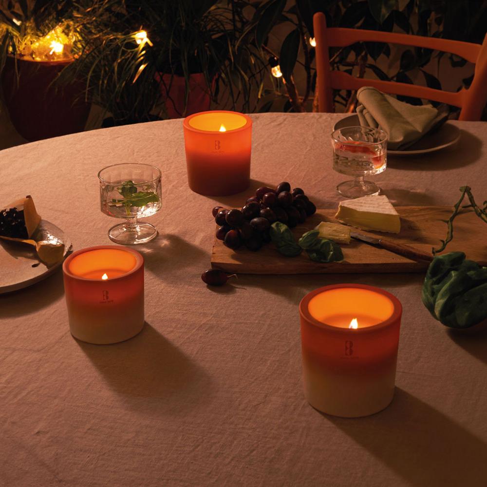 Drei brennende Outdoor Kerzen in orange auf einem gemütlich beleuchteten Tisch, auf dem außerdem ein Brett mit Käse und Trauben auf einer rustikalen Tischdecke stehen.