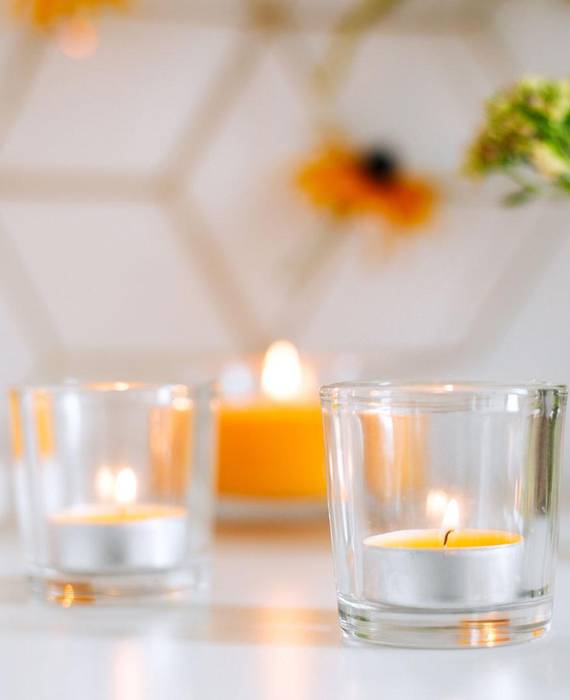 Im Vordergrund steht ein brennendes, gelbes Duft Teelicht in einem Glashalter. Im Hintergrund sieht man weitere gelbe Kerzen brennen, sowie eine Vase mit Blumen.