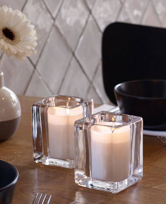 Elegant gedeckter Holztisch mit schwarzem Geschirr und einer runden Vase mit zwei großen, weißen Blüten. In der Mitte des Tisches stehen zwei weiße, brennende Relight Kerzen in einem eckigen Glashalter.