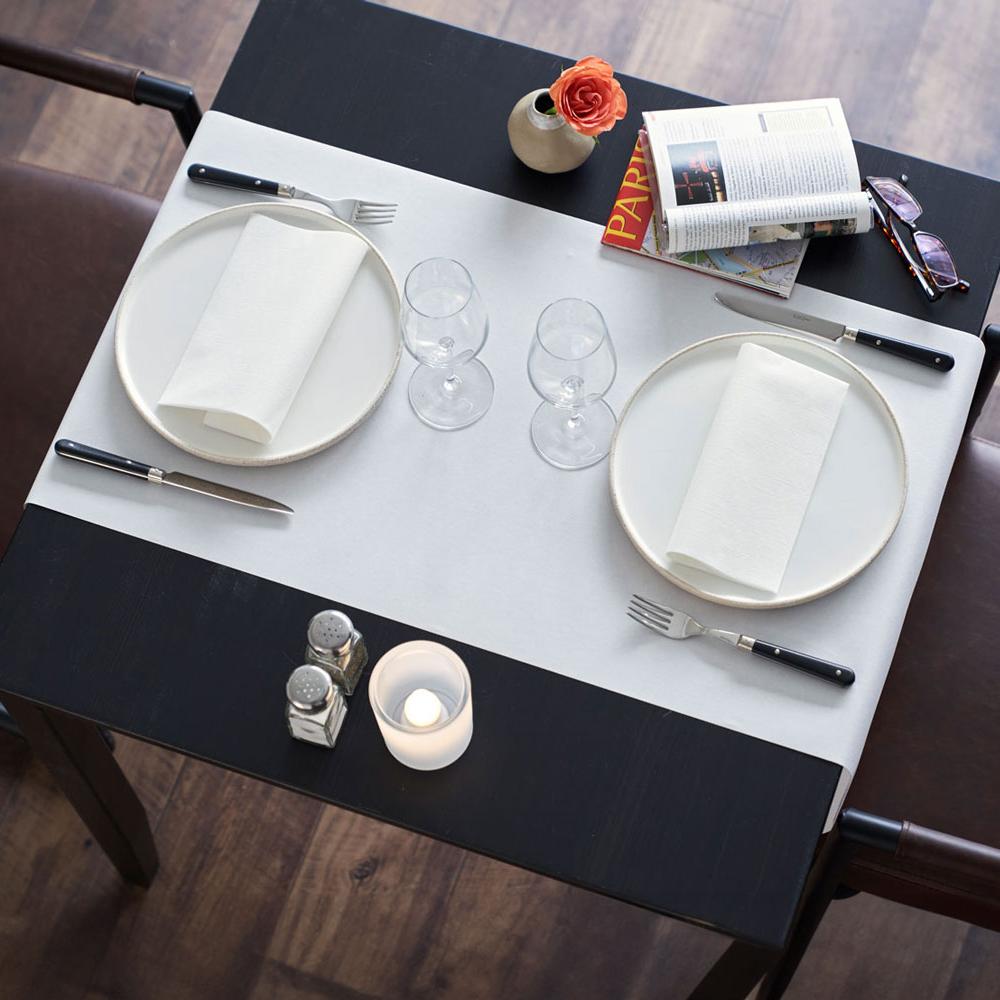 Blick von oben auf einen schlicht und elegant gedeckten Tisch, an dem zwei braune Lederstühle stehen. Unter den Tellern und Weingläsern liegt eine weiße Tischdecke auf dem schwarzen Tisch. Auf den Tellern liegen zwei weiße, gefaltete Servietten.