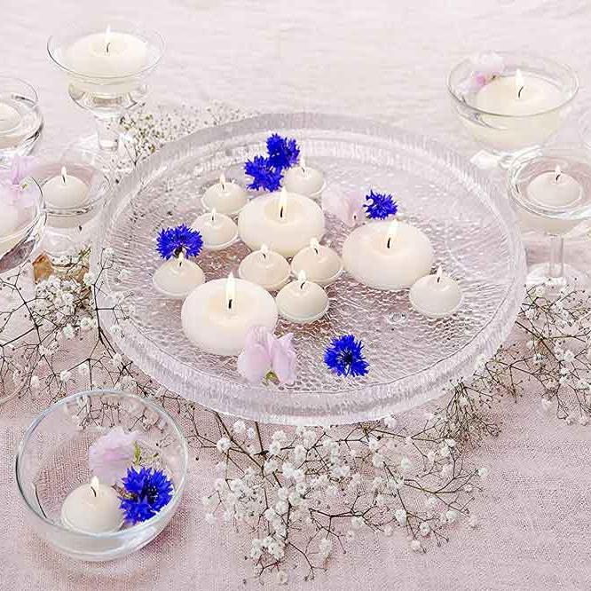 Elfenbeinfarbene Schwimmkerzen und Maxi-Schwimmkerzen in einer Wasserschale und in mit Wasser gefüllten Weingläsern, auf einem mit weißen und blauen Blüten dekoriertem Tisch.