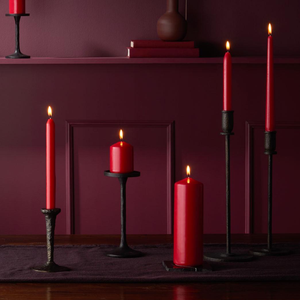 Mehrere rote Stumpenkerzen und Spitzkerzen in verschiedenen Größen, platziert in schwarzen Kerzenhaltern. Dekoriert auf einem dunklen Holztisch mit weinrotem Läufer, vor einer weinroten, holzgetäfelten Wand. 